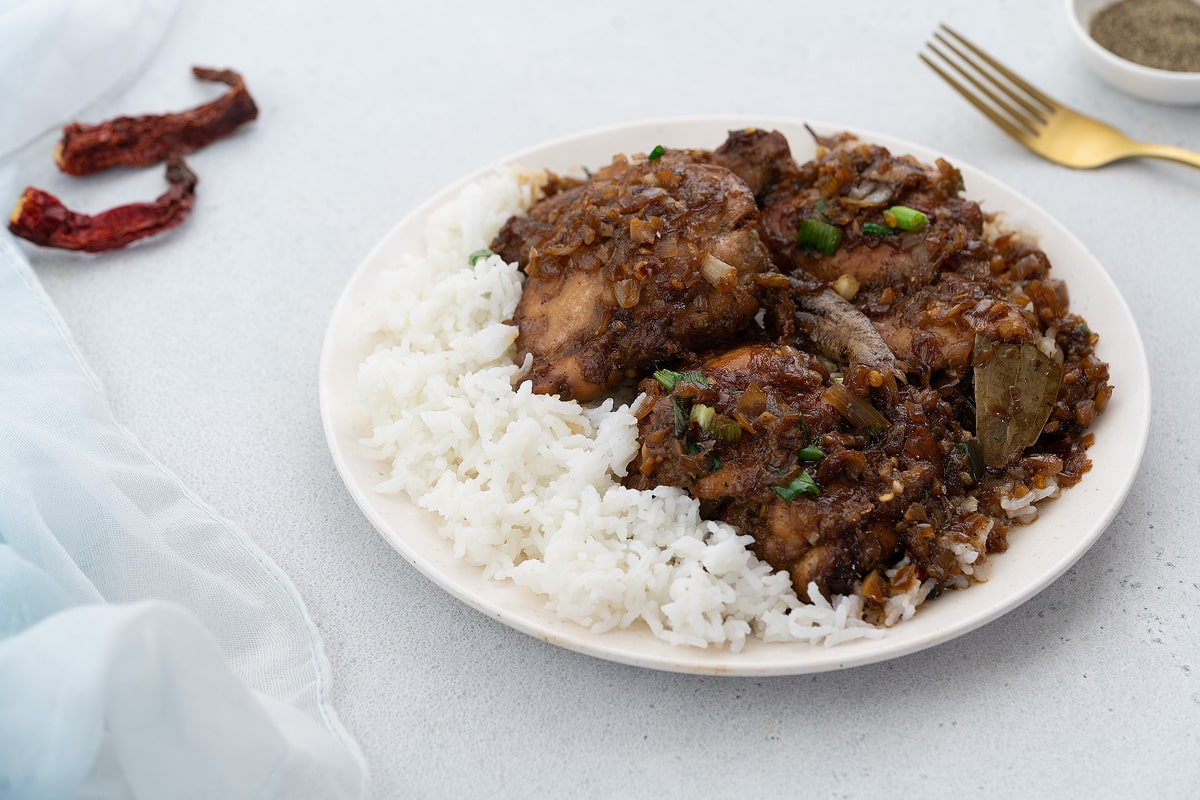 Filipino chicken adobo served on a white plate with a side of rice, accompanied by a golden fork, black pepper on a small plate and chili, all arranged on a white table.
