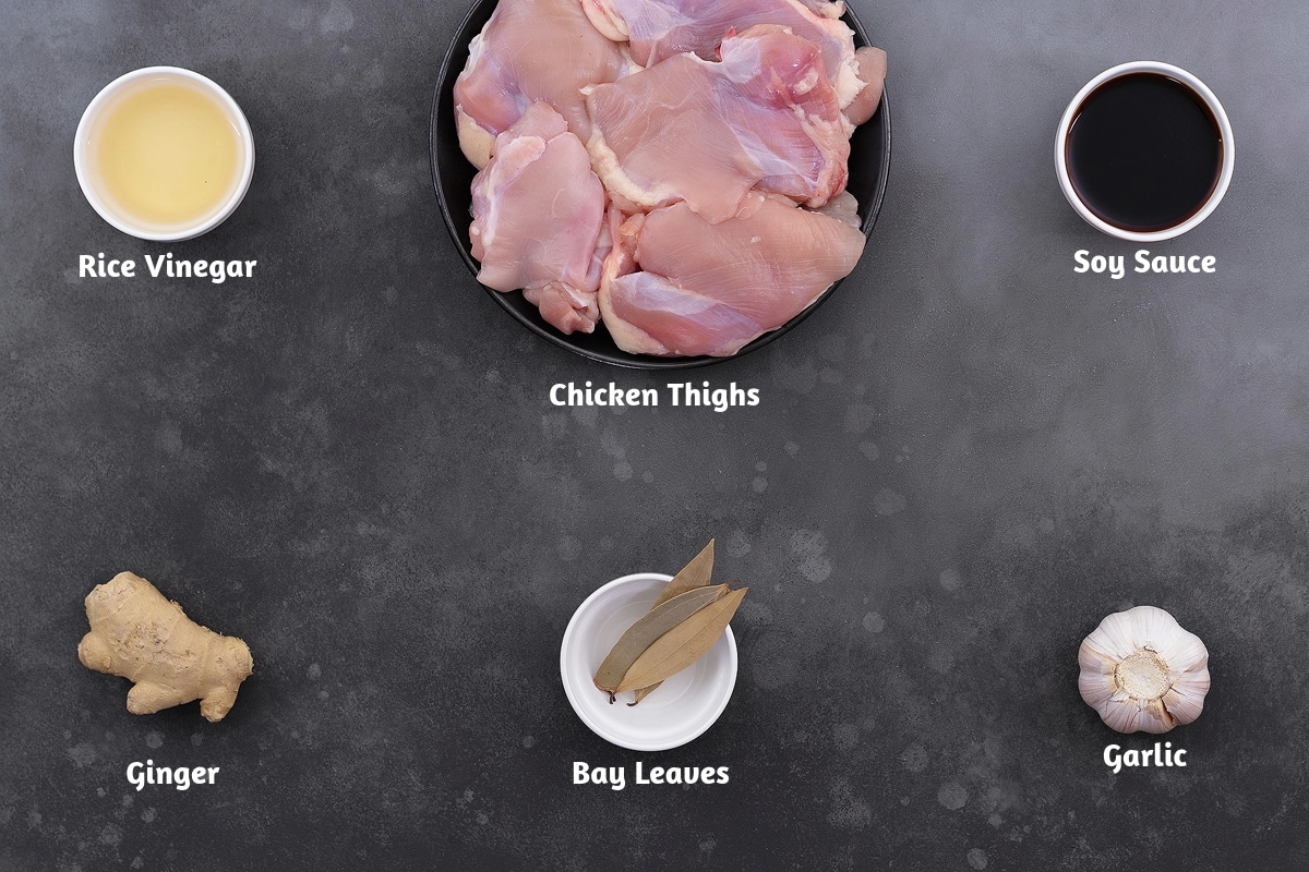 Rice vinegar, chicken thighs, and soy sauce, alongside ginger, bay leaves, and garlic, arranged on a grey table as ingredients for Filipino chicken adobo.