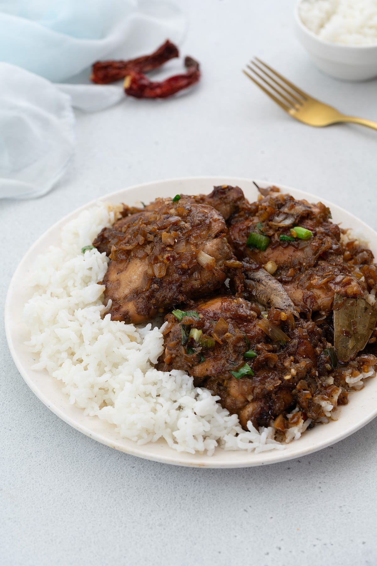 Filipino chicken adobo served on a white plate with a side of rice, accompanied by a golden fork, a cup of rice, and chili, all arranged on a white table.
