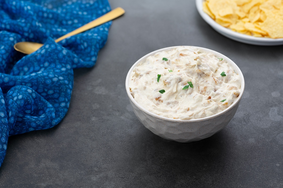 Homemade French onion dip served in a white bowl on a gray table, accompanied by chips, a golden fork, and a blue towel arranged nearby.