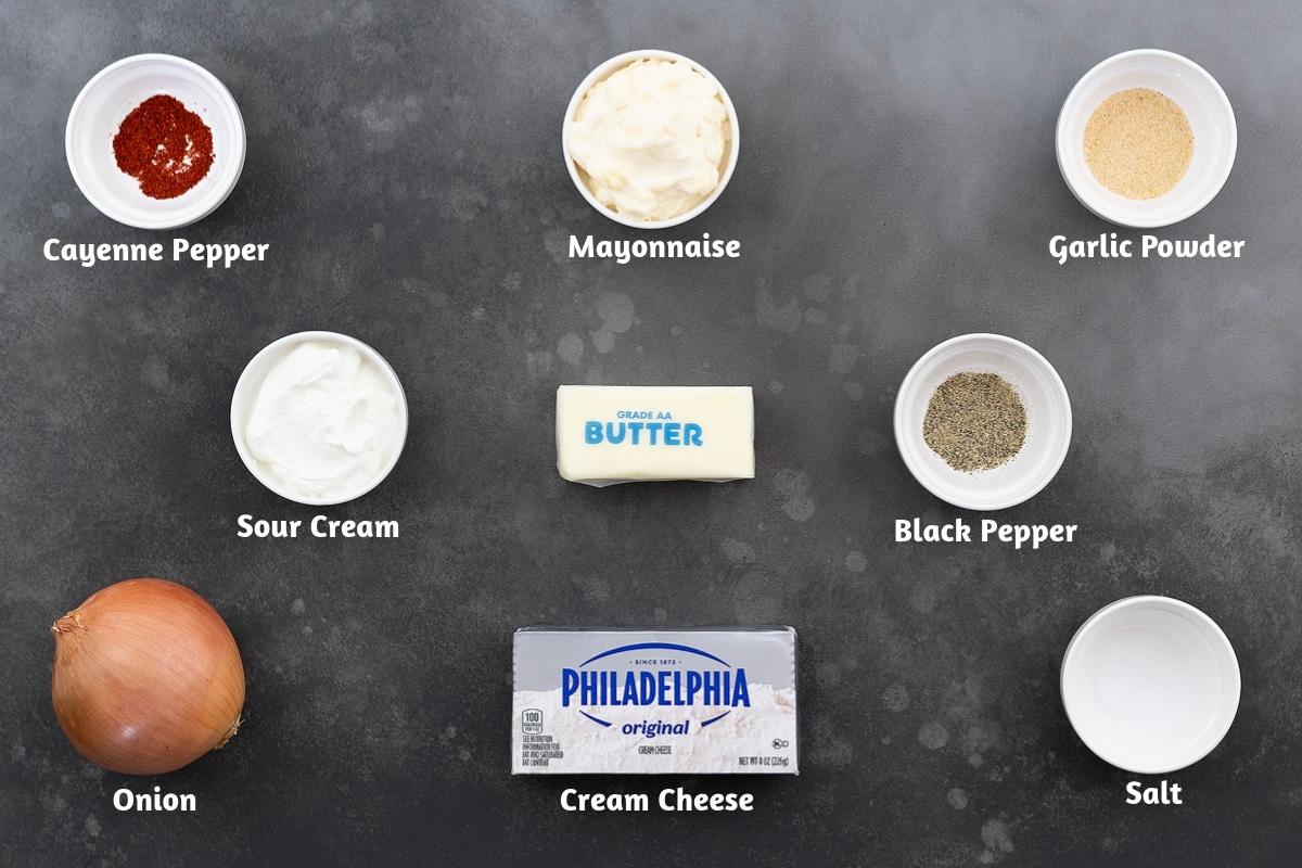 Ingredients for French onion dip arranged on a grey table: cayenne pepper, mayonnaise, garlic powder; sour cream, butter, black pepper; onion, cream cheese, and salt.