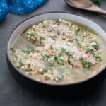 Chicken marinating in Greek-style marinade in a brown bowl, set on a grey table. Nearby are a wooden spoon, a sprig of parsley, and a blue towel.