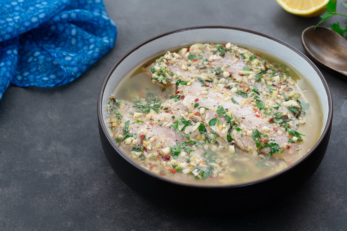 Chicken marinating in Greek-style marinade in a brown bowl, set on a grey table. Nearby are a wooden spoon, a sprig of parsley, a blue towel, and a half-cut lemon.