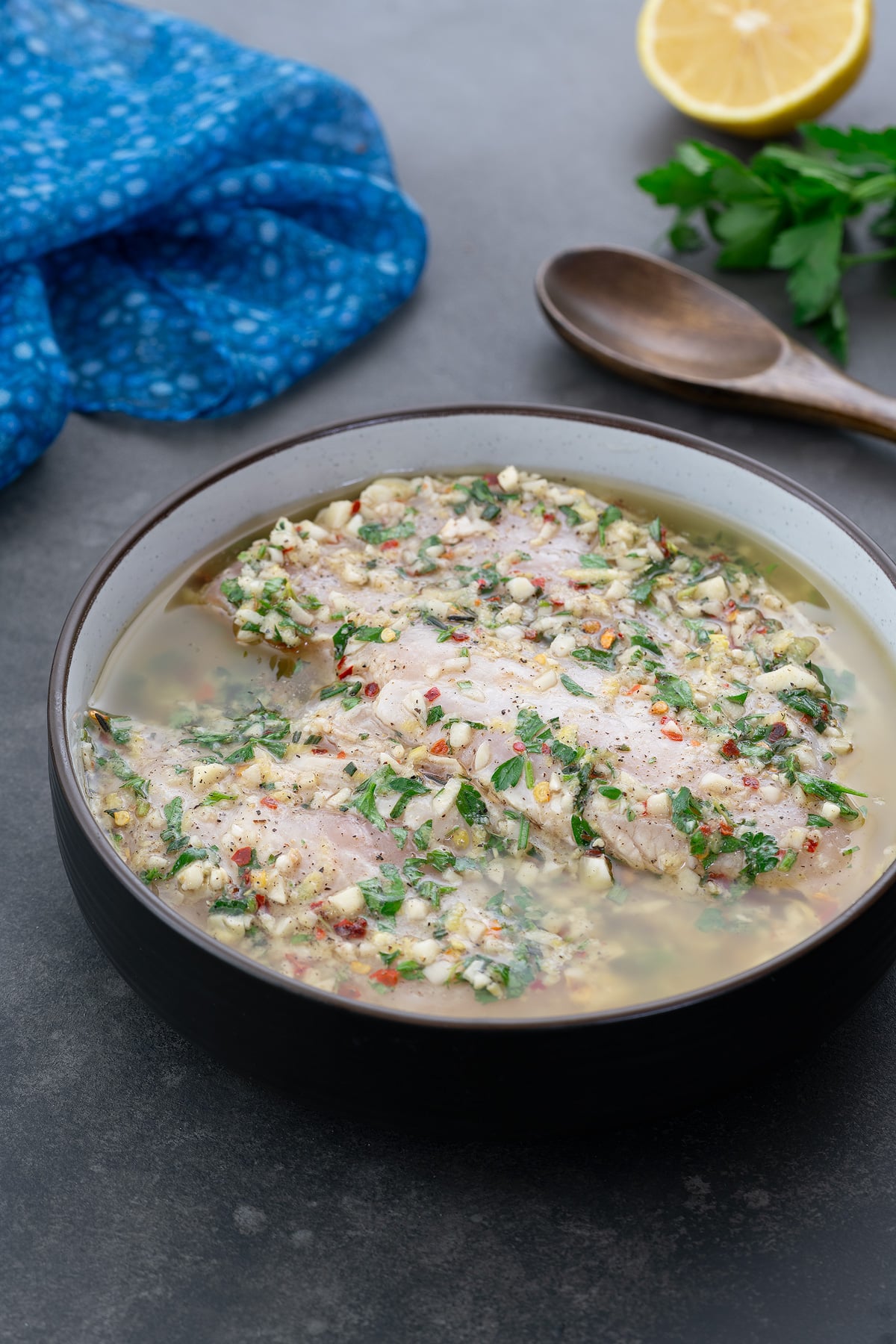 Chicken marinating in Greek-style marinade in a brown bowl, set on a grey table. Nearby are a wooden spoon, a sprig of parsley, a blue towel, and a half-cut lemon.