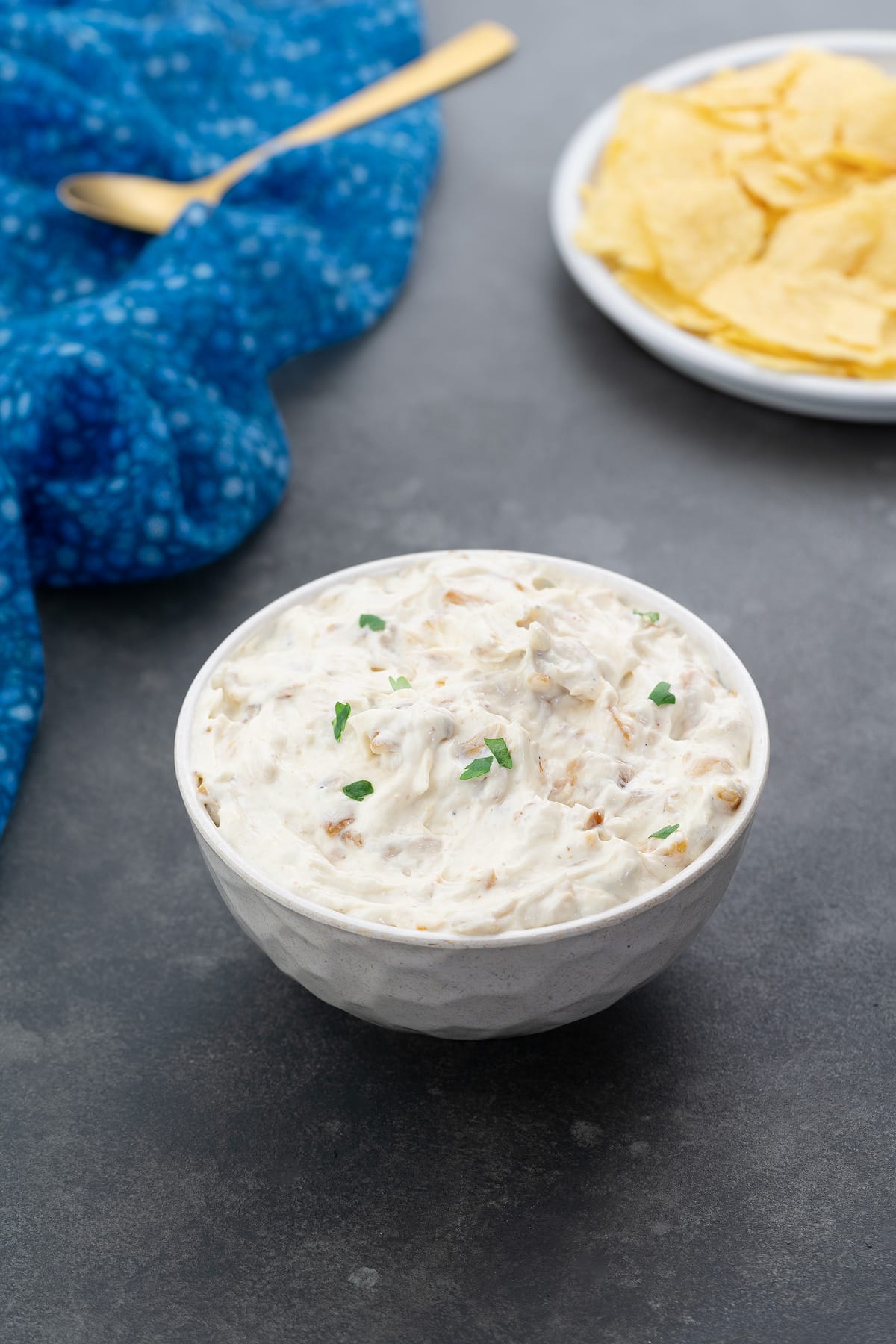 Homemade French onion dip served in a white bowl on a gray table, accompanied by chips, a golden fork, and a blue towel arranged nearby.