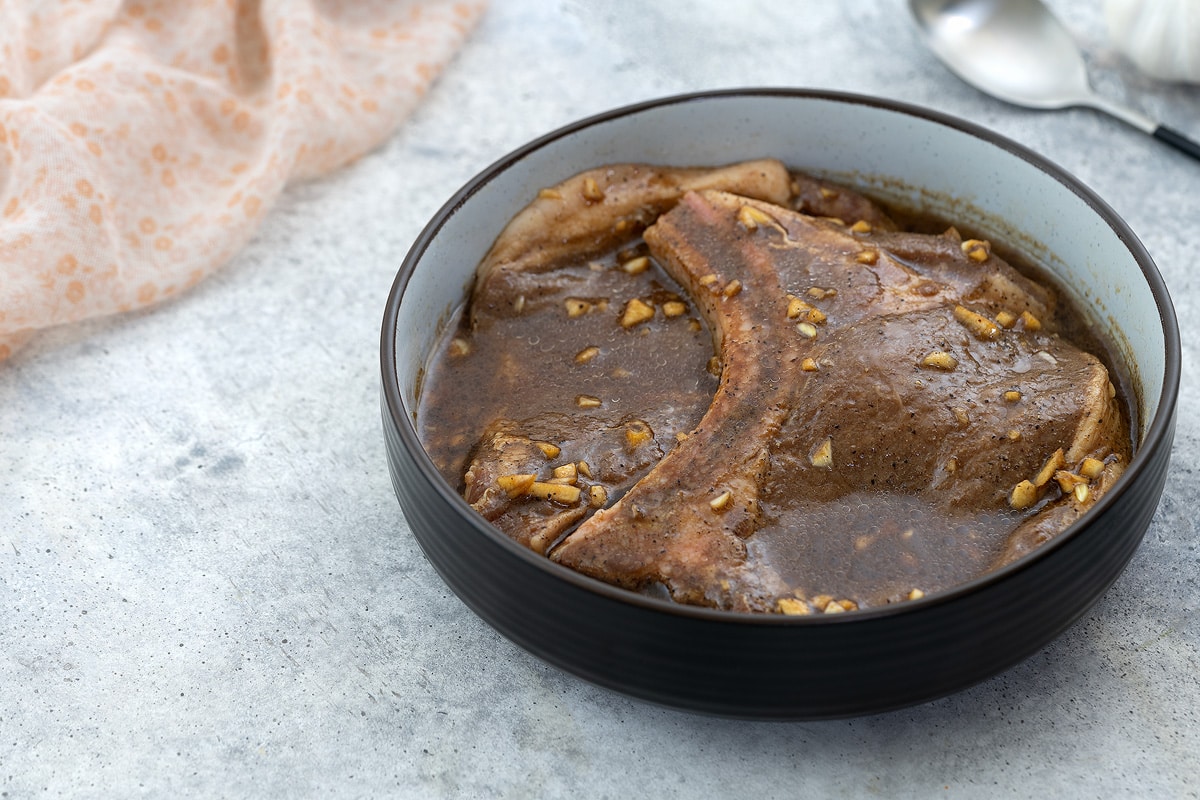 Pork chop marinade in a brown bowl, situated on a white table. A spoon, a bottle of chili flakes, and a towel are neatly arranged around the bowl.