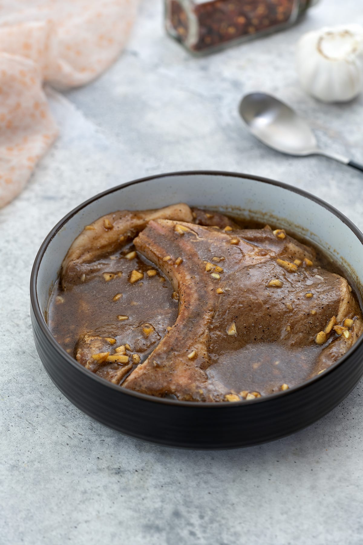 Pork chop marinade in a brown bowl, situated on a white table. A spoon, a bottle of chili flakes, and a towel are neatly arranged around the bowl.