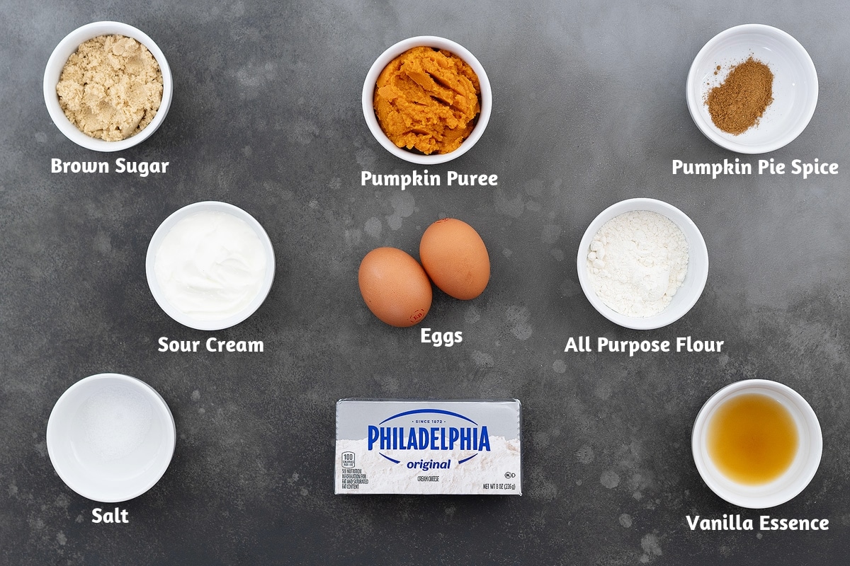 Ingredients for pumpkin cheesecake displayed on a grey table, including brown sugar, pumpkin puree, pumpkin pie spice, sour cream, eggs, all-purpose flour, salt, cream cheese, and vanilla essence.