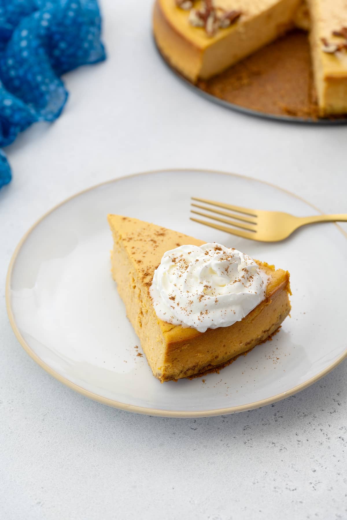Slice of pumpkin cheesecake topped with cream and cinnamon, served on a white plate with a gold fork, accompanied by a blue towel.