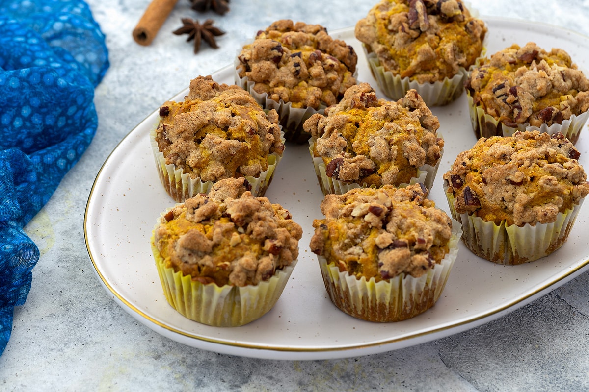 Oval-shaped plate with pumpkin muffins on a white table, accompanied by star anise, a cinnamon stick, and a blue towel nearby.