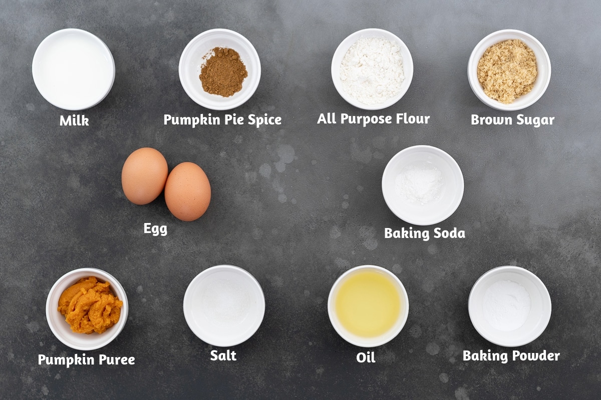 Pumpkin muffin batter ingredients on a grey table, including milk, pumpkin pie spice, all-purpose flour, brown sugar, egg, baking soda, pumpkin puree, salt, oil, and baking powder.