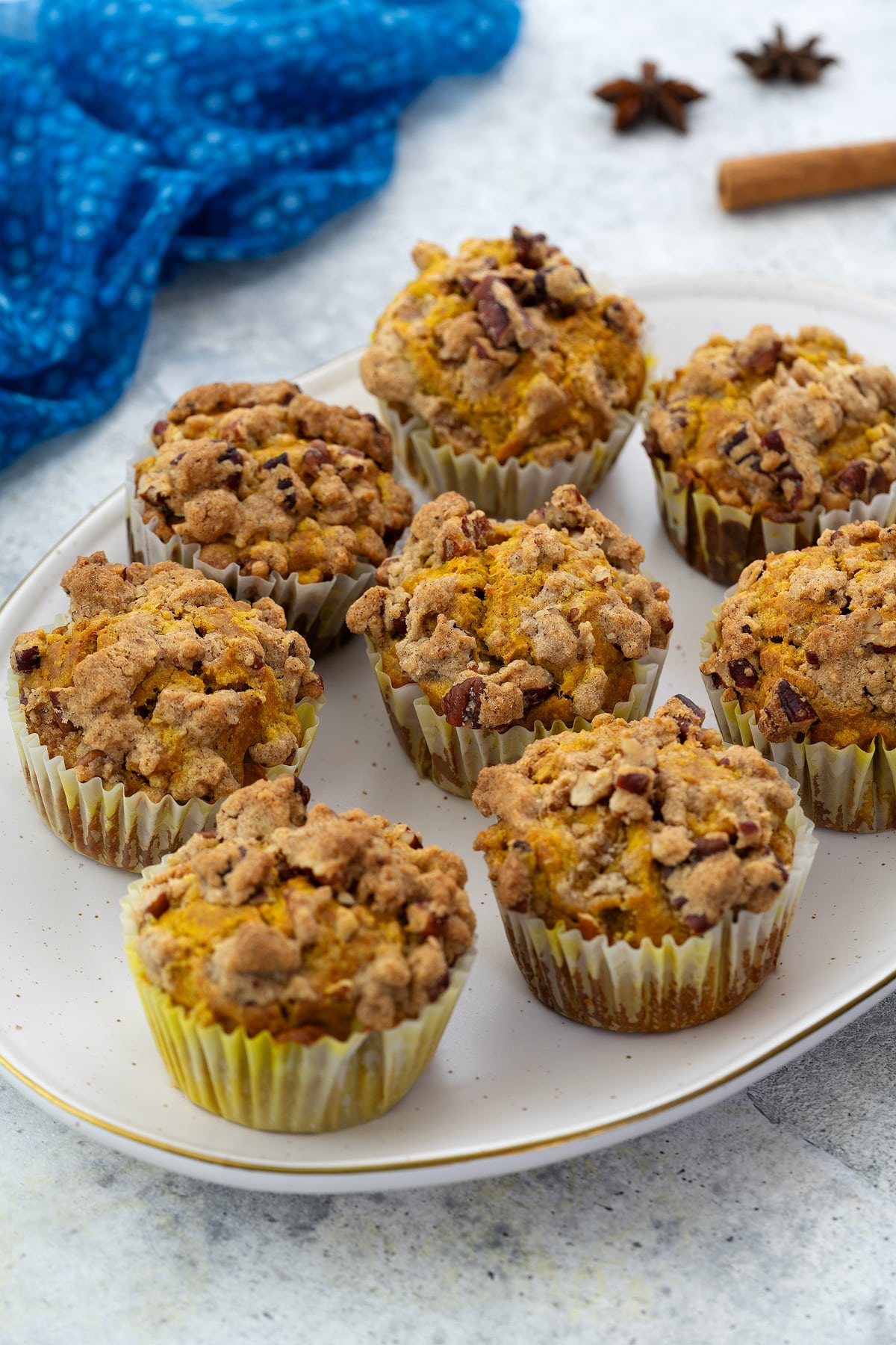 Oval-shaped plate with pumpkin muffins on a white table, accompanied by star anise, a cinnamon stick, and a blue towel nearby.