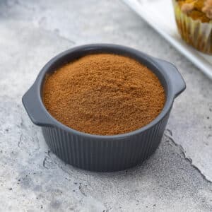 Pumpkin pie spice in a grey bowl on a white table, with a pumpkin muffin arranged around it.