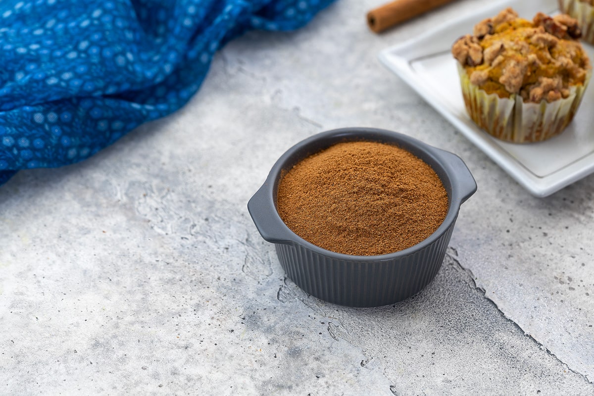 Pumpkin pie spice in a grey bowl on a white table, with a pumpkin muffin, cinnamon stick, and a blue towel arranged around it.