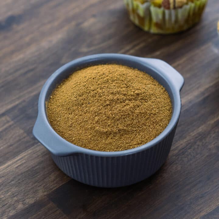 Pumpkin Pie Spice mix in a small grey bowl along with some pumpkin muffins nearby.