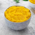 A white bowl of pumpkin puree sits on a white table, surrounded by a cup of puree, a golden fork, and greens.