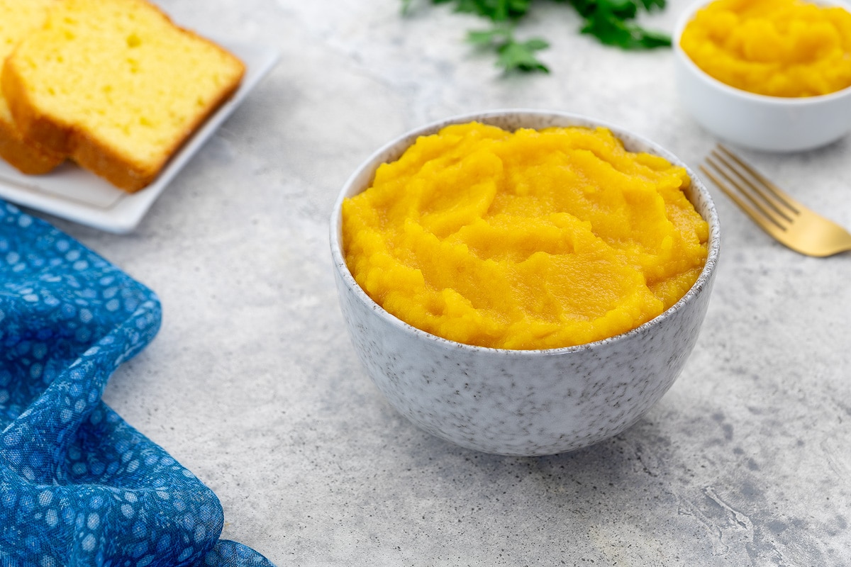 A white bowl of pumpkin puree sits on a white table, surrounded by a cup of puree, pumpkin bread, a golden fork, and greens.