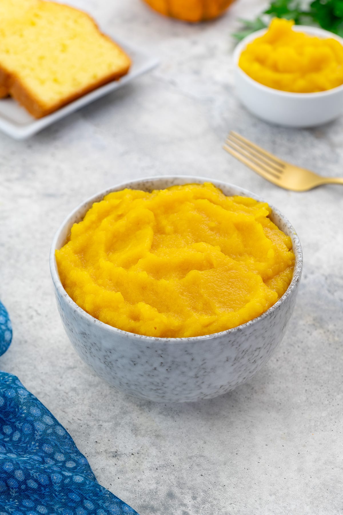 A white bowl of pumpkin puree sits on a white table, surrounded by a cup of puree, pumpkin bread, a golden fork, and greens.