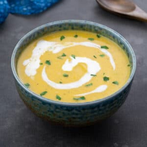 Homemade pumpkin soup served in a green patterned bowl on a gray table. A wooden spoon, and a blue towel are neatly arranged around the bowl.