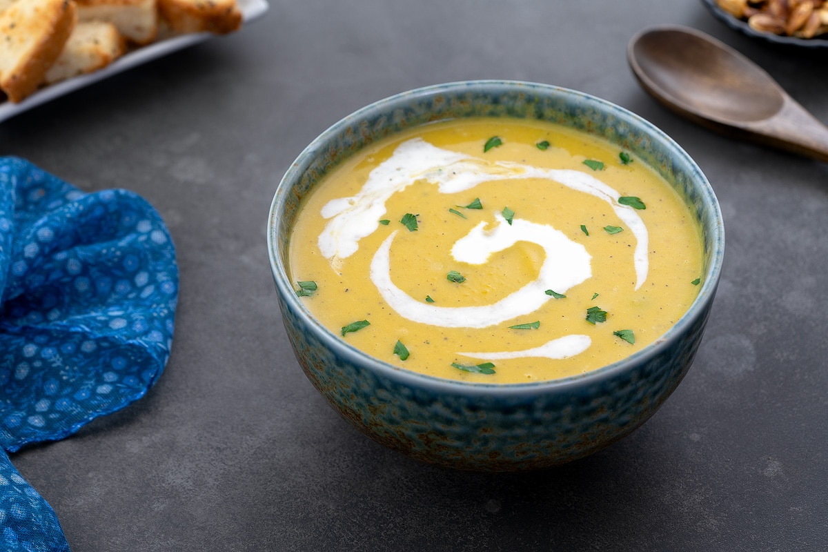 Homemade pumpkin soup served in a green patterned bowl on a gray table. A wooden spoon, croutons, and a blue towel are neatly arranged around the bowl.