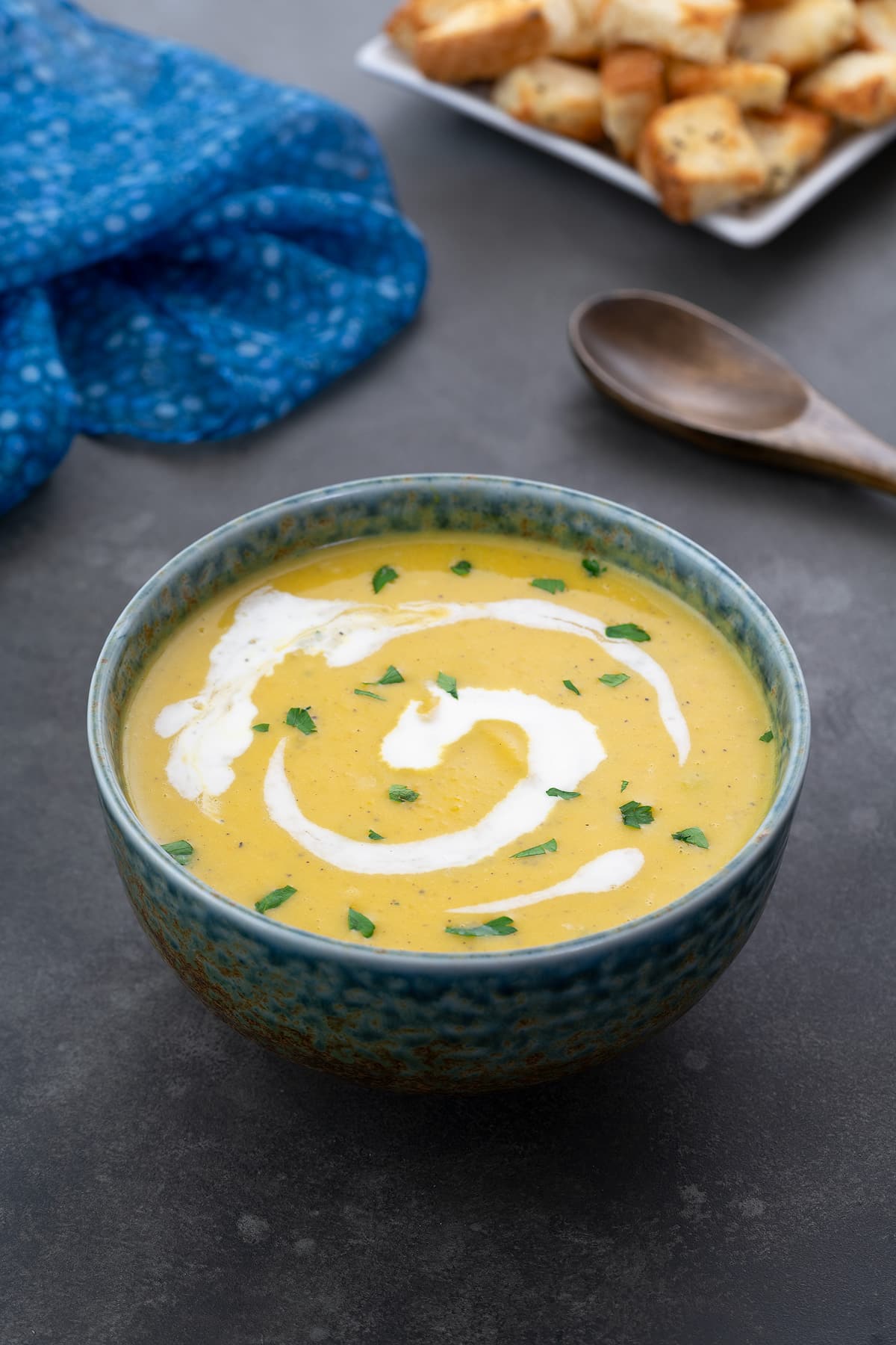 Homemade pumpkin soup served in a green patterned bowl on a gray table. A wooden spoon, croutons, and a blue towel are neatly arranged around the bowl.