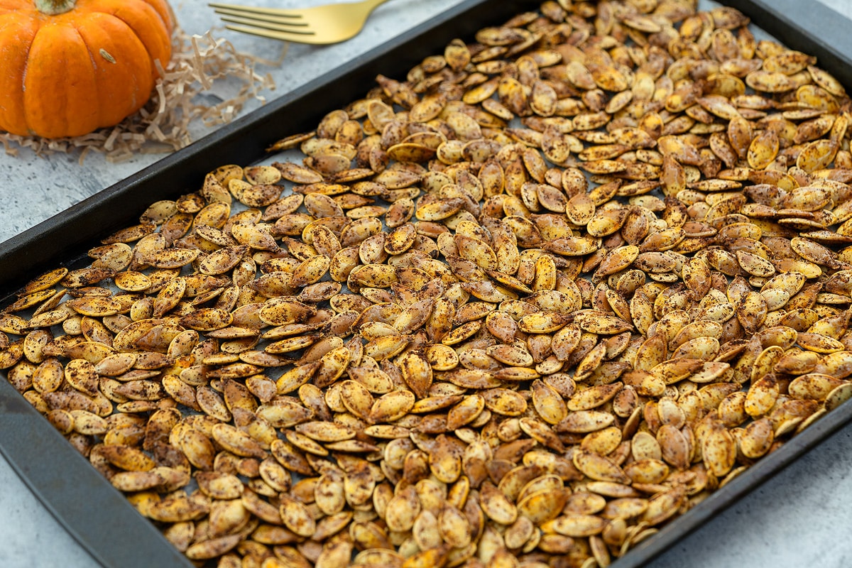 Roasted pumpkin seeds on a baking tray, placed on a white table with a golden fork and a small pumpkin arranged beside it.