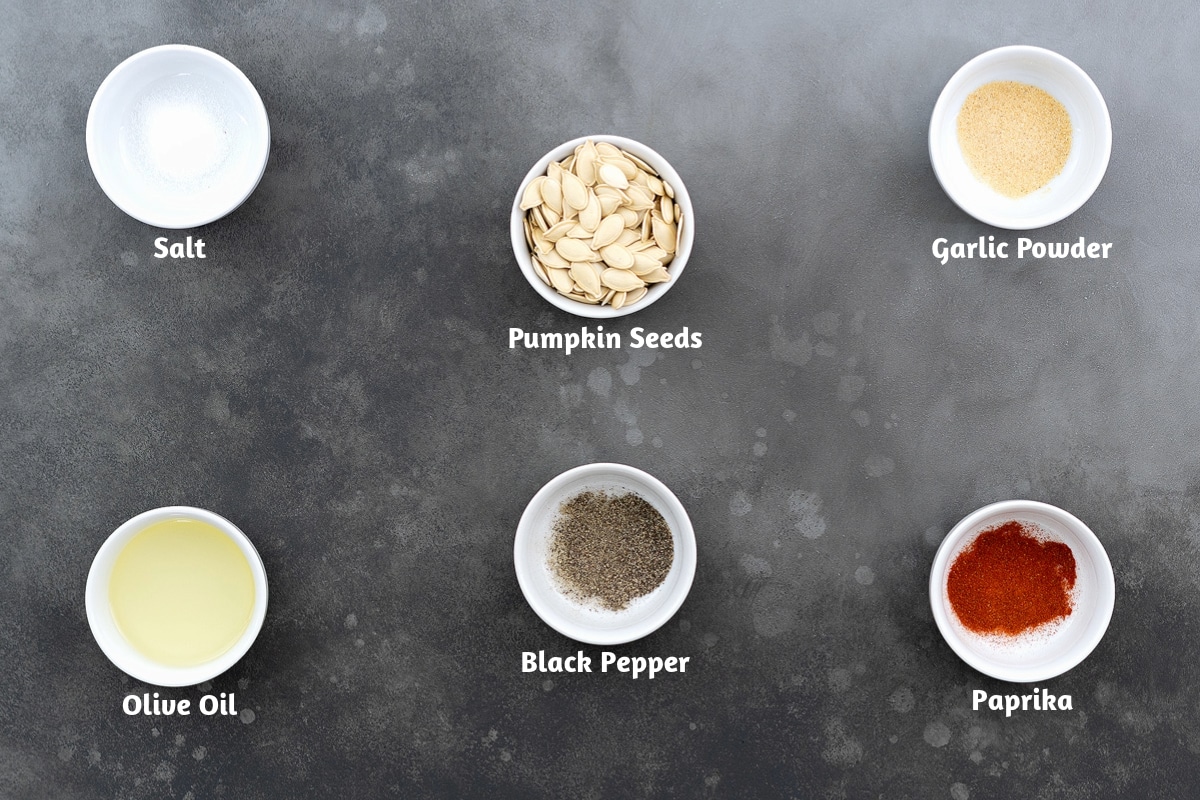 Ingredients for roasted pumpkin seeds arranged on a gray table, including salt, pumpkin seeds, garlic powder, olive oil, black pepper, and paprika.