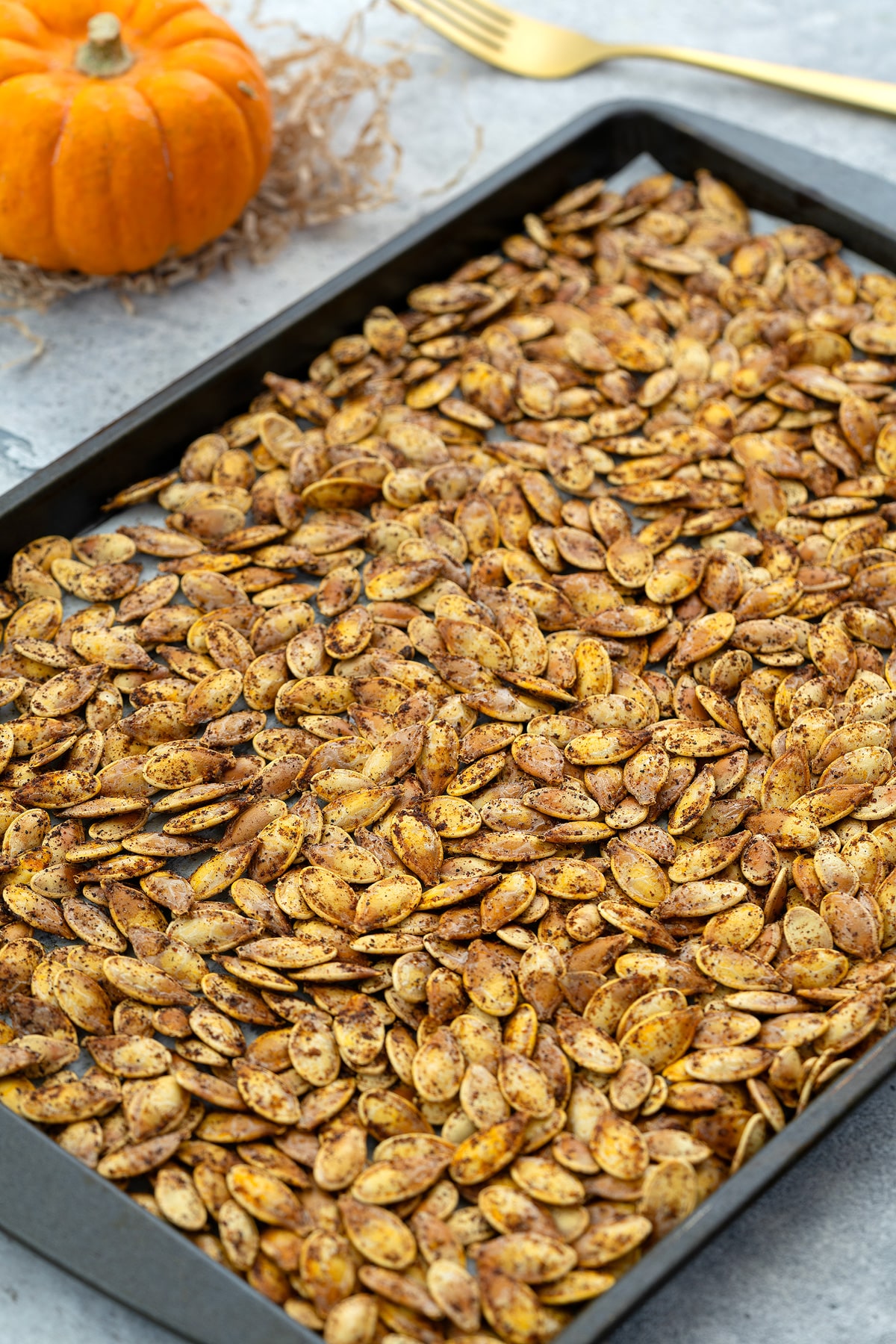 Roasted pumpkin seeds on a baking tray, placed on a white table with a golden fork and a small pumpkin arranged beside it.