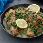 Chicken Piccata in a bowl, topped with lemon slices, butter sauce, greens, and capers, on a gray table.