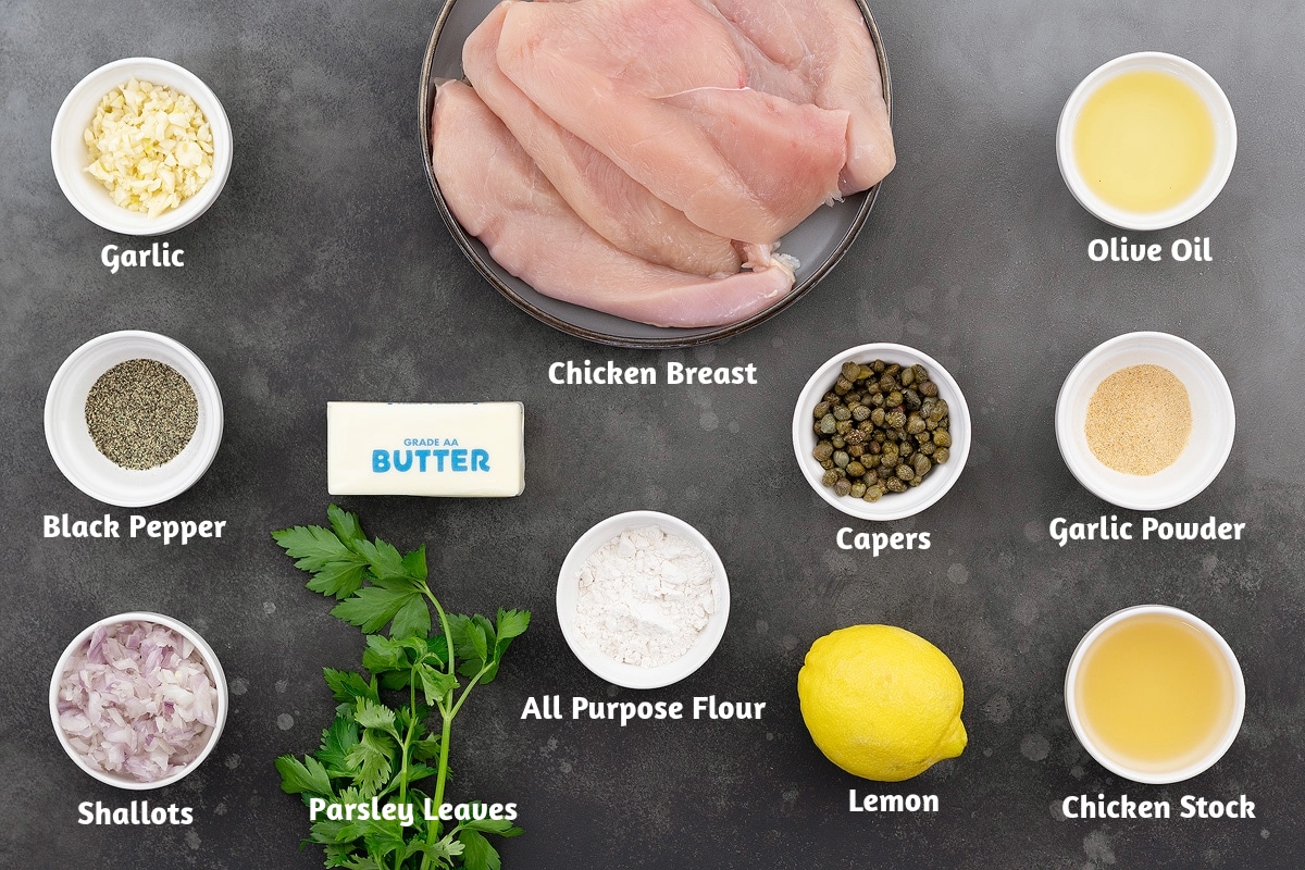 Ingredients for Chicken Piccata on a gray table: garlic, chicken breast, olive oil, black pepper, butter, capers, garlic powder, shallots, parsley, flour, lemon, and chicken stock.