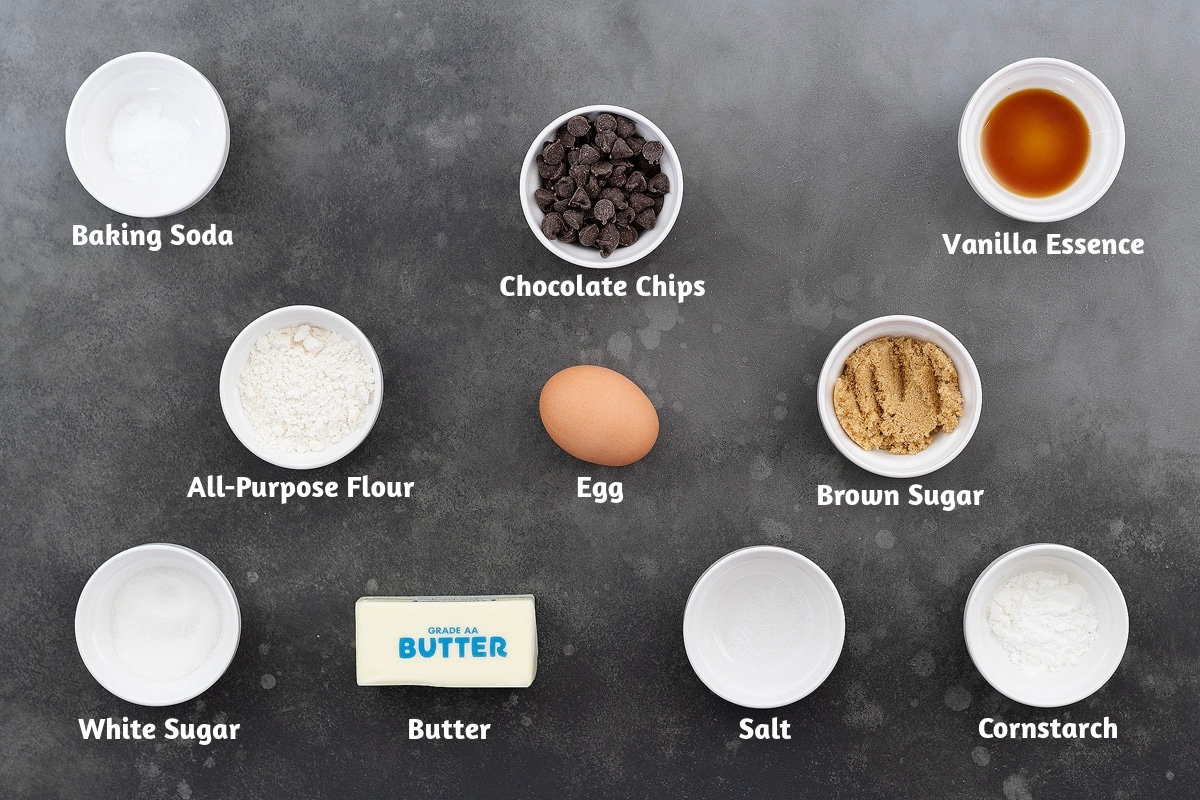 Ingredients for chocolate chip cookies - baking soda, chocolate chips, vanilla essence, all-purpose flour, egg, brown sugar, white sugar, butter, salt, and cornstarch - neatly arranged on a grey table.