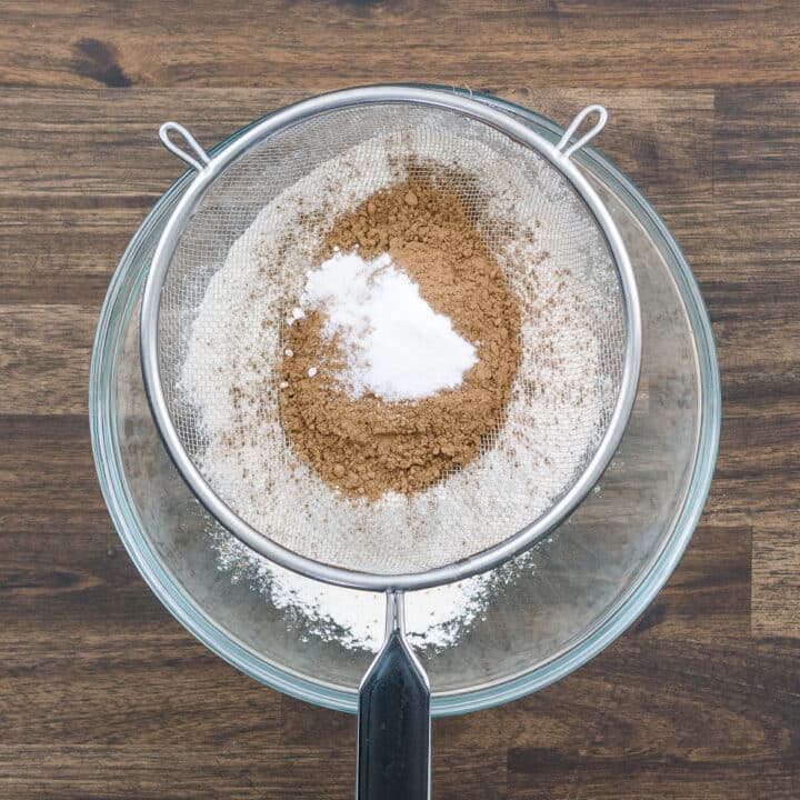 A bowl with flour, cocoa, baking soda, and salt sifted together using a fine mesh strainer.
