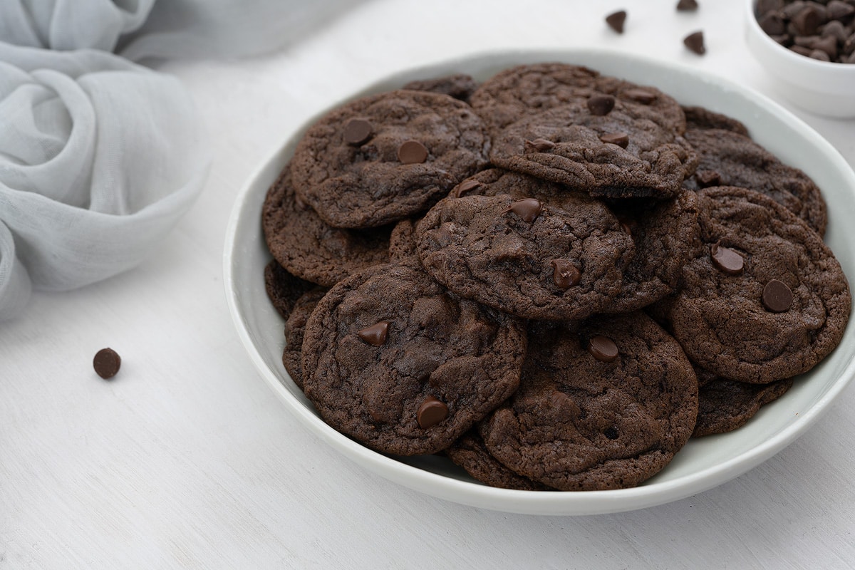 Delicious homemade double chocolate chip cookies displayed on a plate, accompanied by a cup filled with extra chocolate chips and a neatly arranged towel in the background.