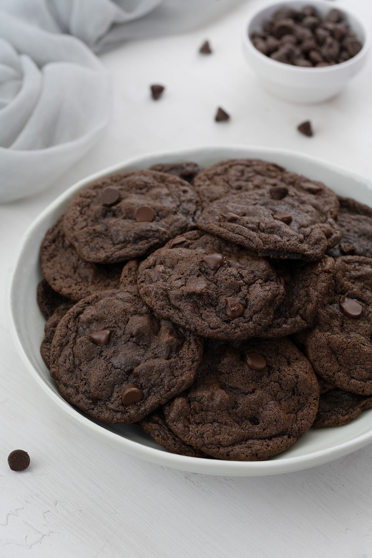 Delicious homemade double chocolate chip cookies displayed on a plate, accompanied by a cup filled with extra chocolate chips and a neatly arranged towel in the background.