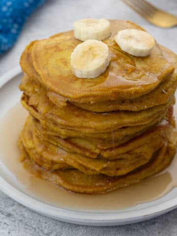 Pumpkin pancake on a white plate, topped with banana slices and drizzled with maple syrup. Beside it is a blue towel, and a golden fork.