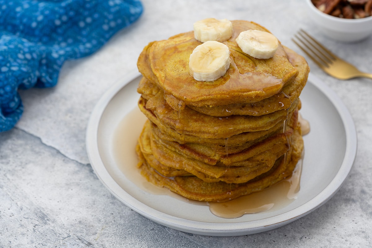 Pumpkin pancake on a white plate, topped with banana slices and drizzled with maple syrup. Beside it is a blue towel, a golden fork, and a cup of nuts.
