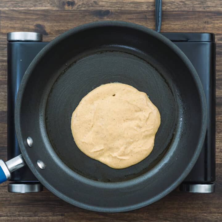 A pan on a stovetop with pumpkin pancake batter cooking, showing bubbles forming on the surface.