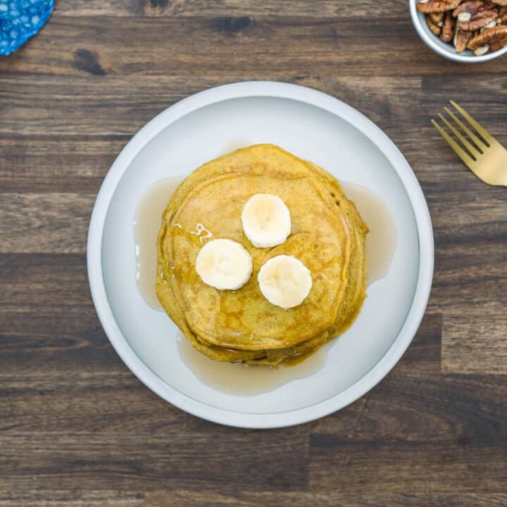 A stack of pumpkin pancakes on a plate, drizzled with maple syrup and topped with banana slices.