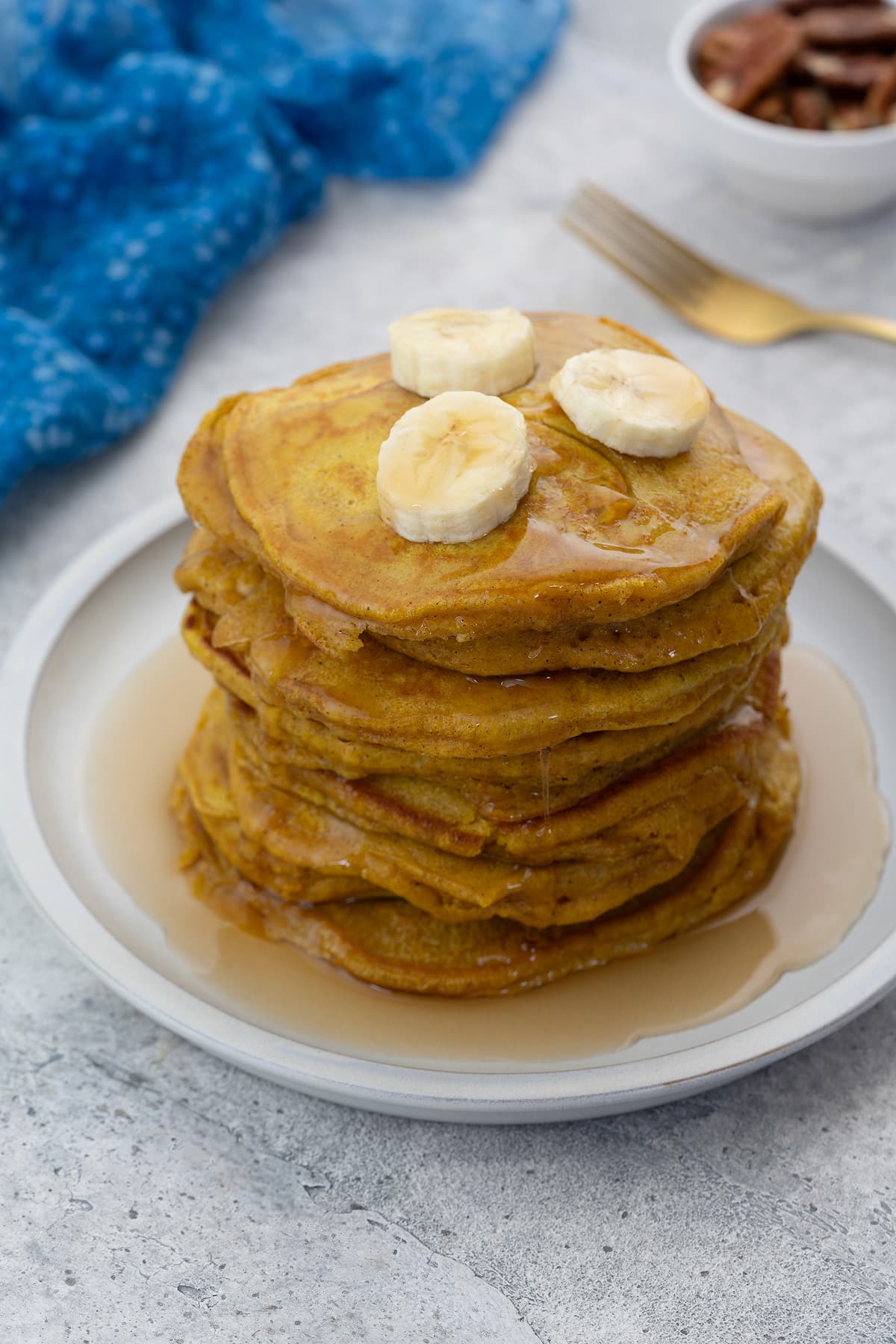Pumpkin pancake on a white plate, topped with banana slices and drizzled with maple syrup. Beside it is a blue towel, a golden fork, and a cup of nuts.