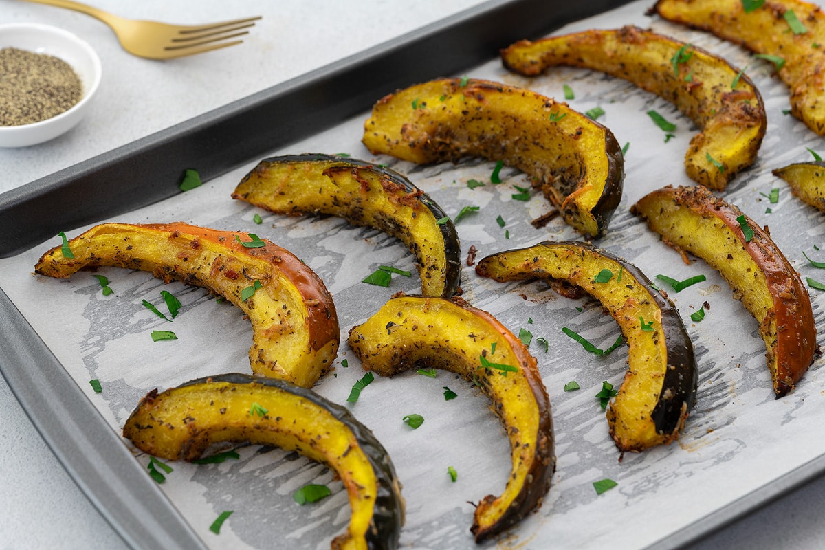 Slices of roasted acorn squash in a baking tray on a white table, accompanied by a golden fork and a small cup of pepper powder arranged nearby.