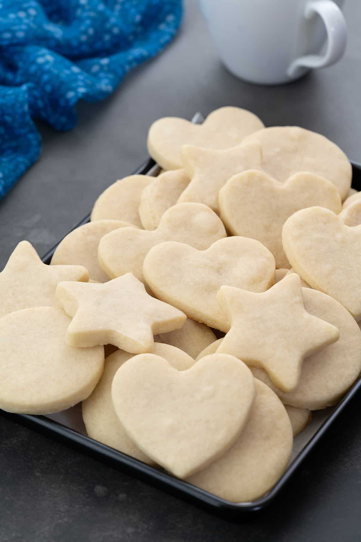 Assorted shortbread cookies in heart, star, and round shapes on a serving tray, accompanied by a coffee cup and a blue towel arranged around it.