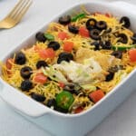 Colorful homemade taco dip in a white baking dish, surrounded by a golden fork.