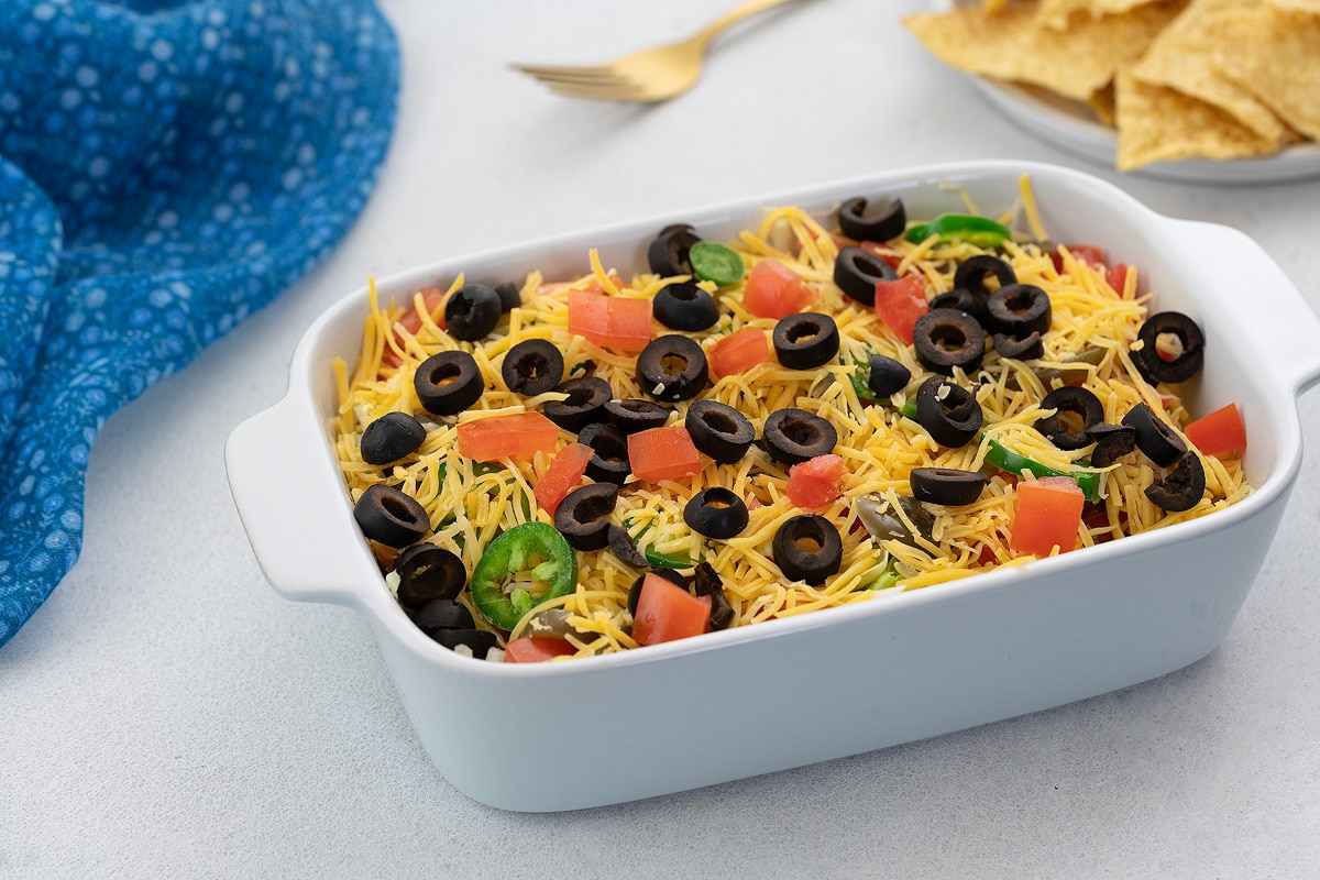 Colorful homemade taco dip in a white baking dish, surrounded by tortilla chips, a towel, and a golden fork.