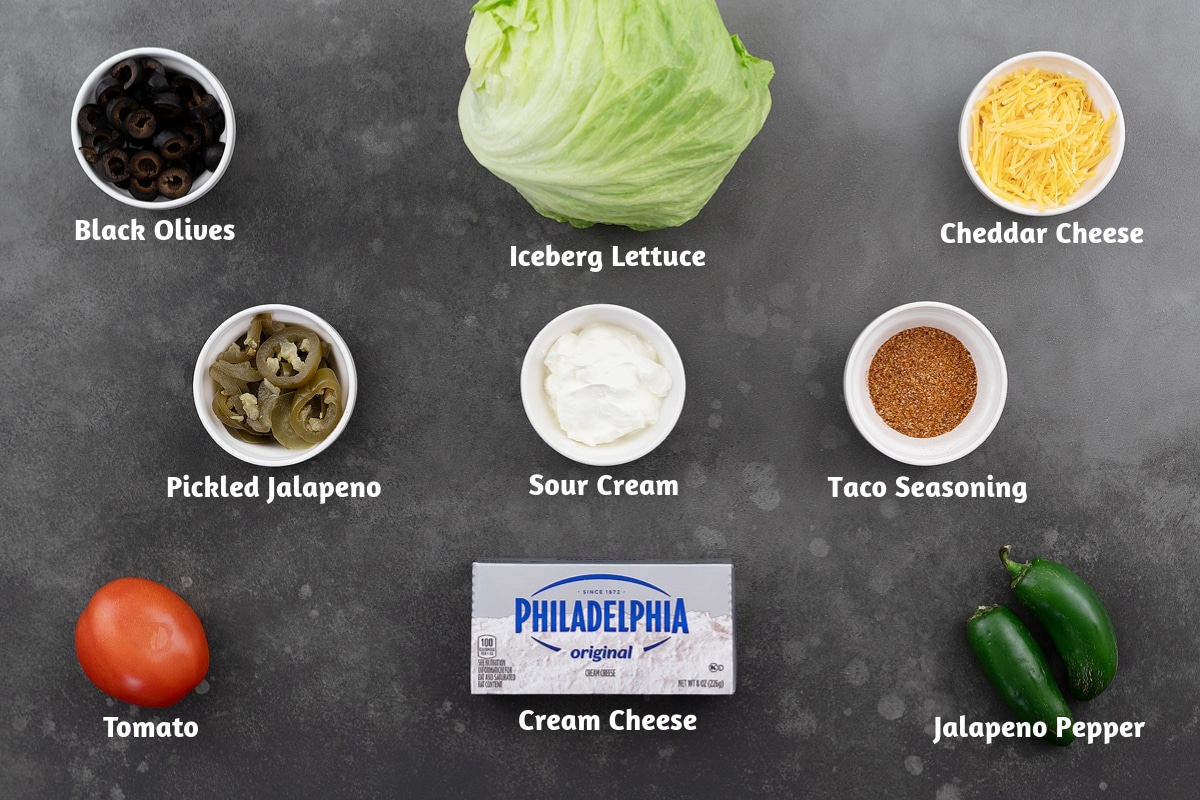 Ingredients for taco dip arranged on a gray table, including black olives, iceberg lettuce, cheddar cheese, pickled jalapeños, sour cream, taco seasoning, tomato, cream cheese, and a jalapeño pepper.