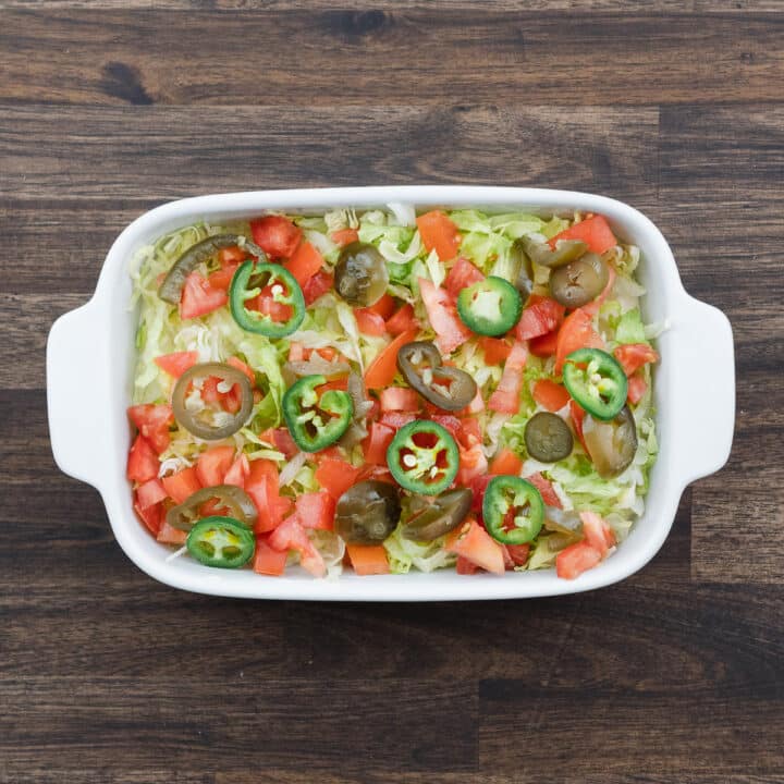 A shallow tray showcasing a creamy base layered with jalapenos, along with lettuce and tomatoes.
