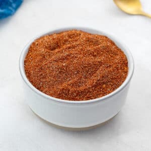 A white bowl filled with homemade BBQ spice rub, set on a white table. Next to it, there's a blue towel and a golden fork artistically arranged.