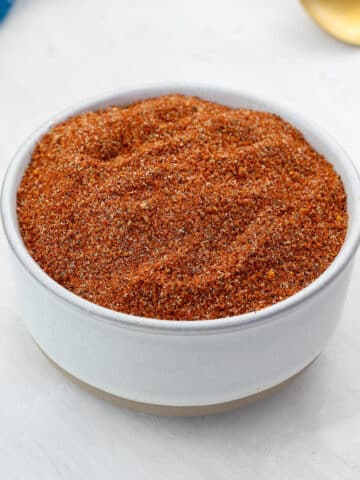A white bowl filled with homemade BBQ spice rub, set on a white table. Next to it, there's a blue towel and a golden fork artistically arranged.