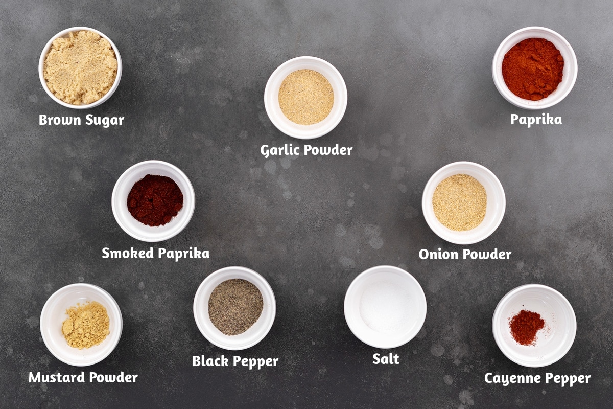 BBQ spice rub ingredients on a gray table: brown sugar, garlic and smoked paprika, onion powder, mustard powder, black pepper, salt, and cayenne pepper.