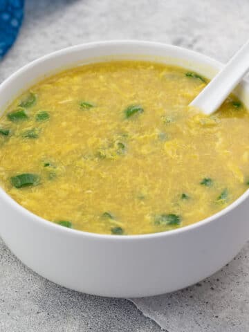 Egg drop soup in a white bowl accompanied by a spoon, set on a gray table.