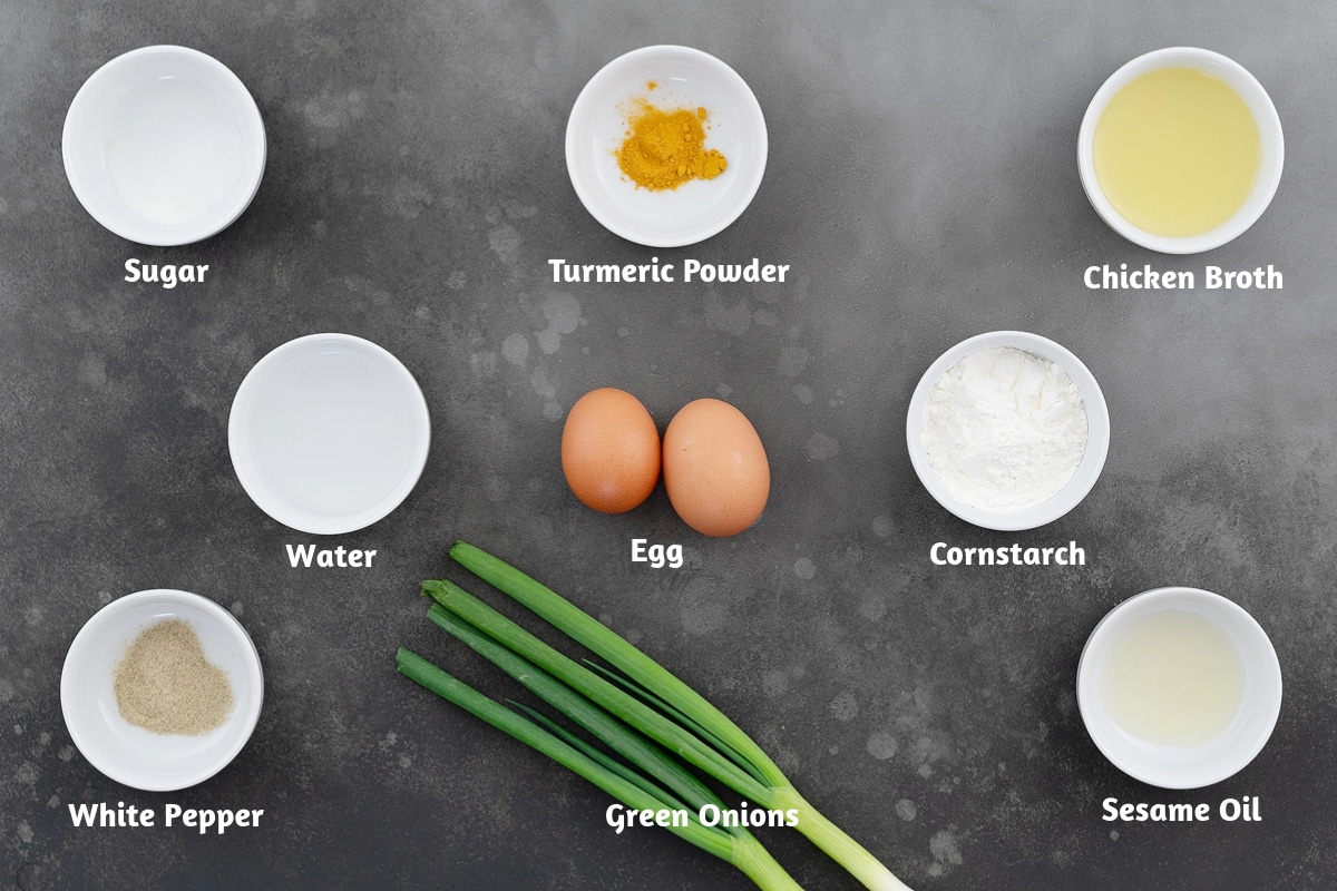 Ingredients for egg drop soup arranged on a gray table, including sugar, turmeric powder, chicken broth, water, an egg, cornstarch, white pepper, chopped green onions, and sesame oil.